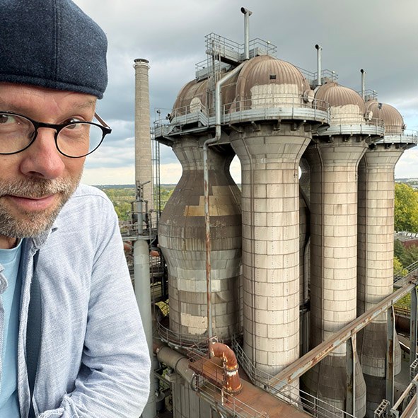 Jaakko Jaskari silmälaseissa ja lippalakissa, taustalla Landschaftspark Duisburgin vanhan masuunin torneja